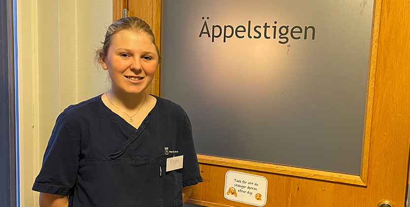 Person med vårdkläder som ler och kollar in i kameran.