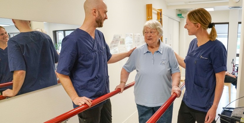 Rehab-lokal med ett par i yrkeskläder som hjälper en dam att träna balans.