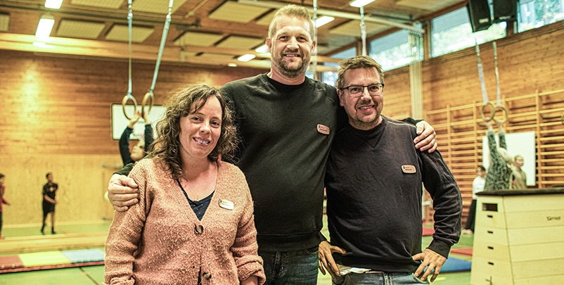 En kvinna och två män i en idrottshall med lekande barn i bakgrunden